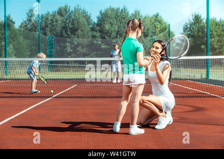 Dark-haired mommy smiling while teaching girl playing tennis Stock Photo