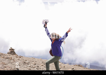 Pretty woman enjoys her extreme trip to the African mountains. Wearing modern blue jacket, grey pants and sunglasees. Long blonde hair waving on wind Stock Photo