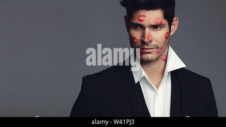 Handsome young man with lipstick marks of kisses on his face. Handsome male model in black blazer looking away while standing against grey background. Stock Photo