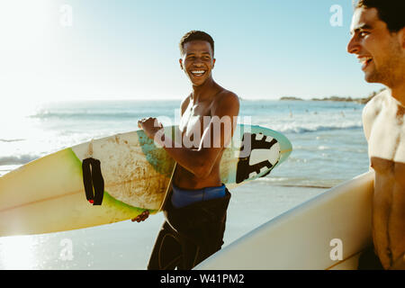 Smiling men walking on the beach carrying surfboards. Male friends on surfing vacation at the sea. Stock Photo