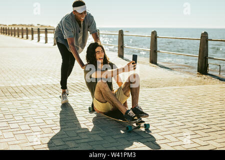 Cheerful friends playing on the skateboard outdoor and taking a selfie. Young guy taking selfie while sitting on skateboard with a friend pushing him. Stock Photo