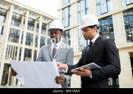 Two engineers in suits and helmets discussing project of new building together while standing in the city with modern building behind their backs Stock Photo