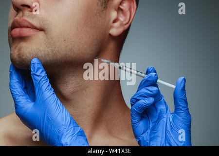 Close-up portrait of young man isolated on grey studio background. Filling botox surgery procedure. Concept of men's health and beauty, cosmetology, self-care, body and skin care. Anti-aging. Stock Photo