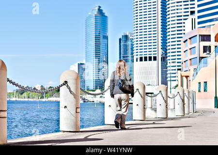 Back view of a woman walking along the footpath with concrete pillar fences beside blue water river and under a sky from Brisbane City, Queensland, Au Stock Photo
