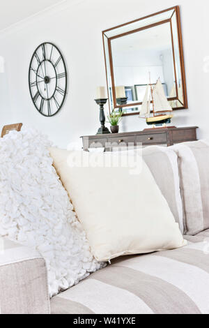 White pillows on chair, luxury looking new sofa set near wooden table, toy ship also candle can see on  tables, clock attached to wall, daytime colorf Stock Photo