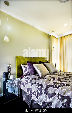Flower designed bed sheet near to shiny pillows in bedroom including a window closed with curtains, there is a small mirror table with fancy vase Stock Photo
