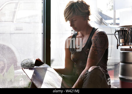 Tattoo girl with gold color short hair takes out the vinyl disk from the album cover and sitting beside a glass window Stock Photo