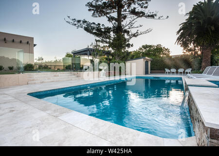 Luxury swimming pool view with blue water poolside with chairs and small fountain from a corner with trees surrounded by a glass wall and trees in the Stock Photo