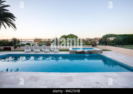 Luxury swimming pool with a small fountain covered with fence beside trees from the garden under blue sky, there are chairs around the waterside Stock Photo