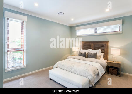 An antique oak wood finish bed set paired with white bed covers, grey throw-pillows and a white bed-foot bench adds to the charm of this classic- cont Stock Photo
