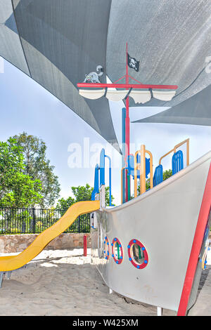 Play equipment with ship design, sands are white color, sky is blue and have clouds, there is a flag on top of ship, green trees and fence can see beh Stock Photo