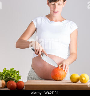 Young pregnant woman preparing vegetables in the kitchen cutting a fresh autumn pumpkin on a wooden chopping board with three fresh lemons alongside f Stock Photo