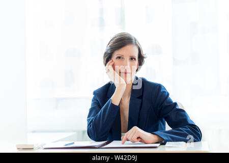 Thoughtful businesswoman staring pensively ahead at the camera with her chin resting on her hand as she sits at a desk in the office working on docume Stock Photo