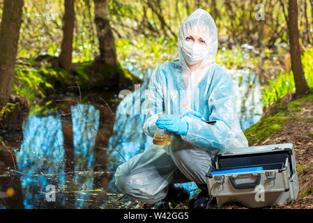 portrait of an ecologist in protective clothing while working, taking water samples from a forest river for analysis Stock Photo