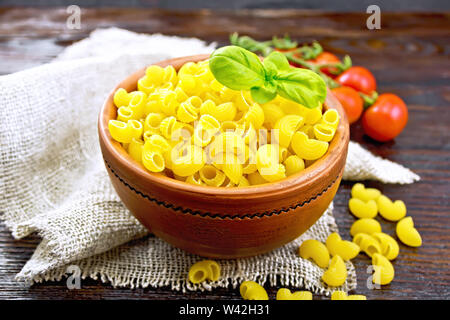 Pasta Elbow with basil in a clay bowl on napkin of burlap, tomatoes on wooden board background Stock Photo