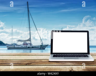 computer laptop with blank white screen on wooden table with yacht boats floating on the sea at background. Travel vacation concept. Stock Photo