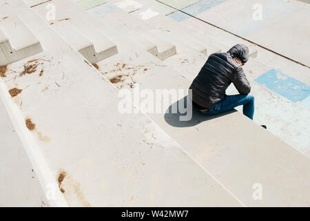 a young caucasian man, seen from behind, wearing jeans and a gray hooded jacket, curled up sitting on an outdoor concrete stairway Stock Photo