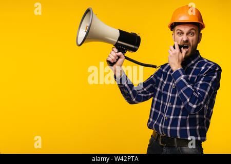 Angry worker man in orange helmet with a megaphone on yellow background - Image Stock Photo