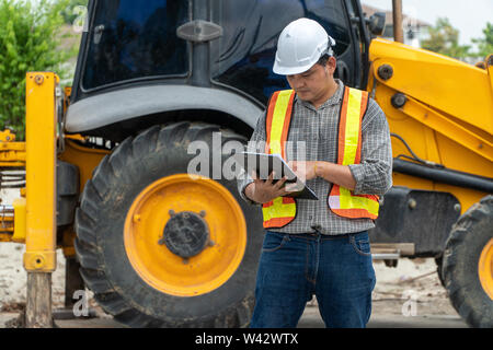 Engineering wearing a white safety helmet standing In front of the backhoe And are using tablet for check the blueprint with construction Stock Photo