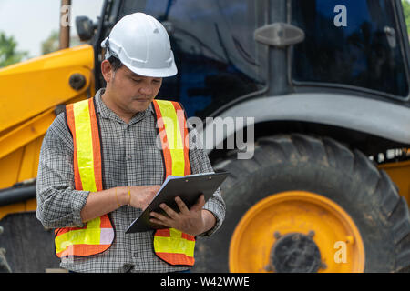 Engineering wearing a white safety helmet standing In front of the backhoe And are using tablet for check the blueprint with construction Stock Photo