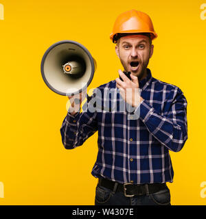 Angry worker man in orange helmet with a megaphone on yellow background - Image Stock Photo