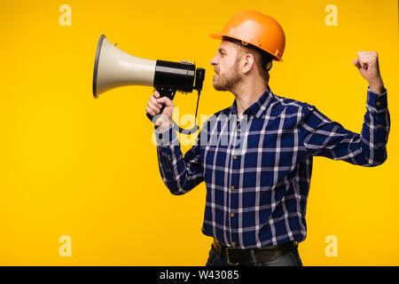 Angry worker man in orange helmet with a megaphone on yellow background - Image Stock Photo