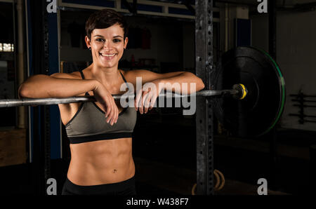 Portrait of an athletic beautiful woman with strong abs in a gym. The short haired female athlete is smiling and leaning against the barbell rack. Stock Photo