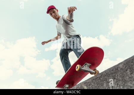 Skateboarder doing a trick at the city's street in sunny day. Young man in sneakers and cap riding and longboarding on the asphalt. Concept of leisure activity, sport, extreme, hobby and motion. Stock Photo