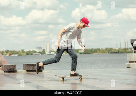 Skateboarder doing a trick at the city's street in sunny day. Young man in sneakers and cap riding and longboarding on the asphalt. Concept of leisure activity, sport, extreme, hobby and motion. Stock Photo