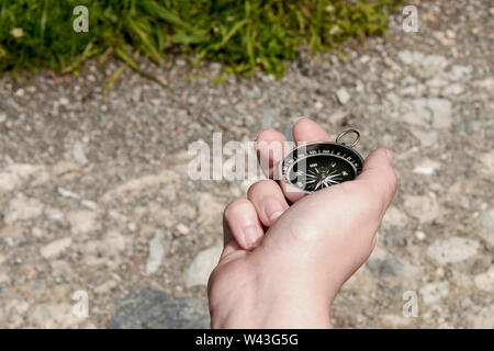 Female hand holding fingers compass pointing north. Stony path with grass on a sunny summer day. Stock Photo