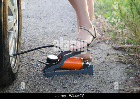 The woman herself pumps up the cars tire with a foot mechanical pump. Feet clad in sandals. The side of the road. Close-up view. Tyre pressure check. Stock Photo