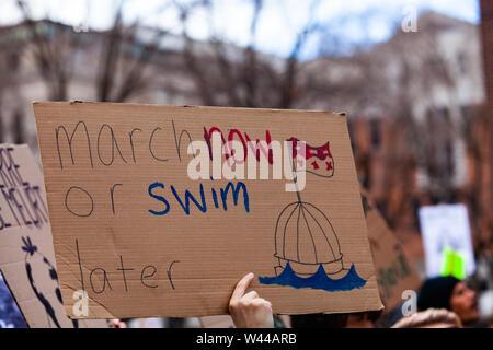 A cardboard sign is viewed close up saying march now or swim later as ecological activists stage a demonstration in a city center.