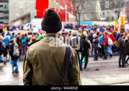 A young man is seen from the back, wearing an olive jacket and wooly hat, as eco-activists march against global warming in a blurry background. Stock Photo