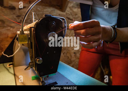 Hands of a young dressmaker are viewed close up, fiddling with red red thread on an industrial sewing machine, preparing to create new garment in workshop. Stock Photo