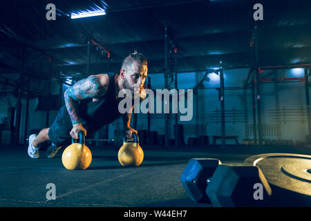 Young healthy man athlete doing push-ups with the weights in the gym. Single male model training and practicing hard. Concept of healthy lifestyle, sport, fitness, bodybuilding, cross fit. Stock Photo