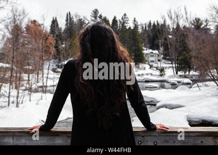 A rear view of a lady with long hair leaning against a wood handrail, contemplating the heavy snowfall and natural winter landscape in the background Stock Photo