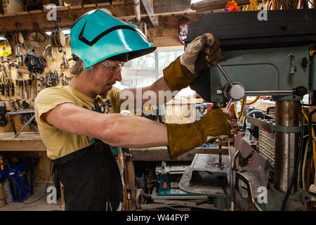 A side profile view of a skilled tradesman operating heavy duty machinery in his workshop. The man uses his arms to operate a drill press on his workt Stock Photo