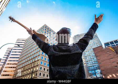 A bohemian man holding a small acoustic guitar is viewed from the back, holding his arms up towards commercial buildings in the city center. Stock Photo