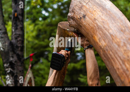 A close up view on the hands of a man using a cordless drill to join a rustic timber structure, construction of a teepee tent as hippies celebrate cultures. Stock Photo