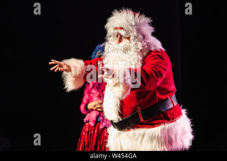 A large man dressed in a Saint Nicholas costume is seen reaching his arms out in front to address youngsters at a christmas festival, against a black background Stock Photo