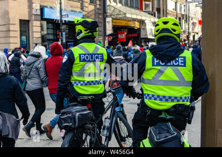 Two on duty police officers wearing high visibility uniforms are seen from behind with pedal bikes, watching people for public safety during a street rally. Stock Photo