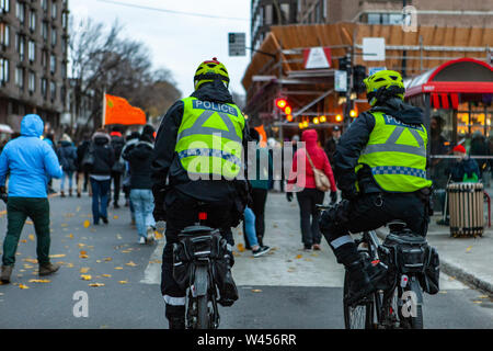 Two policemen are seen from the rear, riding bicycles during a demonstration by environmentalists, protestors are seen in the background on an urban street Stock Photo