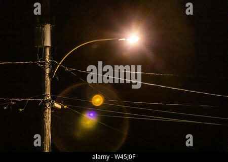 A low angle view of a lamppost lit-up over an urban street by night, overhead cables attach to a utility pole, against a black sky with colorful lens flare. Stock Photo
