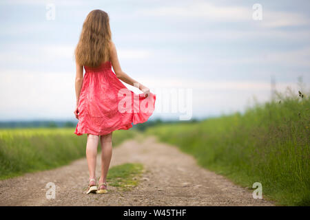 Back view of young romantic slim woman in red dress with long hair walking by ground road along green field on sunny summer day on blue sky copy space Stock Photo