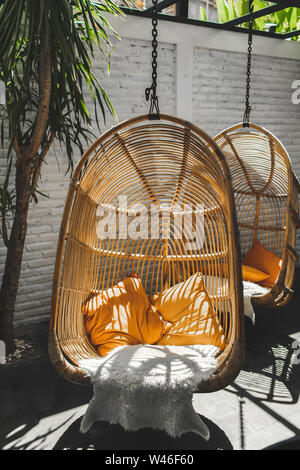 Wicker rattan hanging chair in loft cafe. Eco friendly furniture style and concept. Orange pillows and soft fur on chair. Hipster cafe. Stock Photo