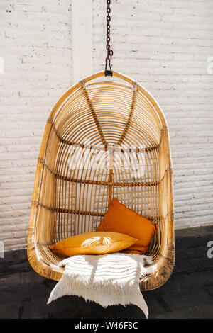 Wicker rattan hanging chair in loft cafe. Eco friendly furniture style and concept. Orange pillows and soft fur on chair. Hipster cafe. Stock Photo