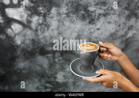 Gray cup of fresh cappuccino in woman hands on background of gray concrete cement textured wall. Trendy hipster cafe design. Stock Photo