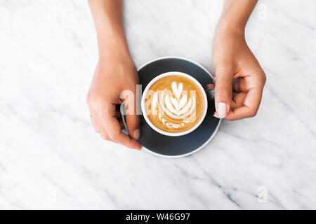 Woman hands holding gray cup of fresh cappuccino with classic latte art on white marble table trendy background. Empty place for text, copy space. Cof Stock Photo