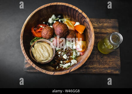 Wooden bowl with falafel balls, hummus and fresh salad. Olive oil in bottle on side. Dark black background, top view. Traditional israeli food. Vegani Stock Photo