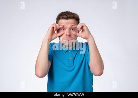 Young man in blue clothes trying to open eyes with fingers, sleepy and tired Stock Photo